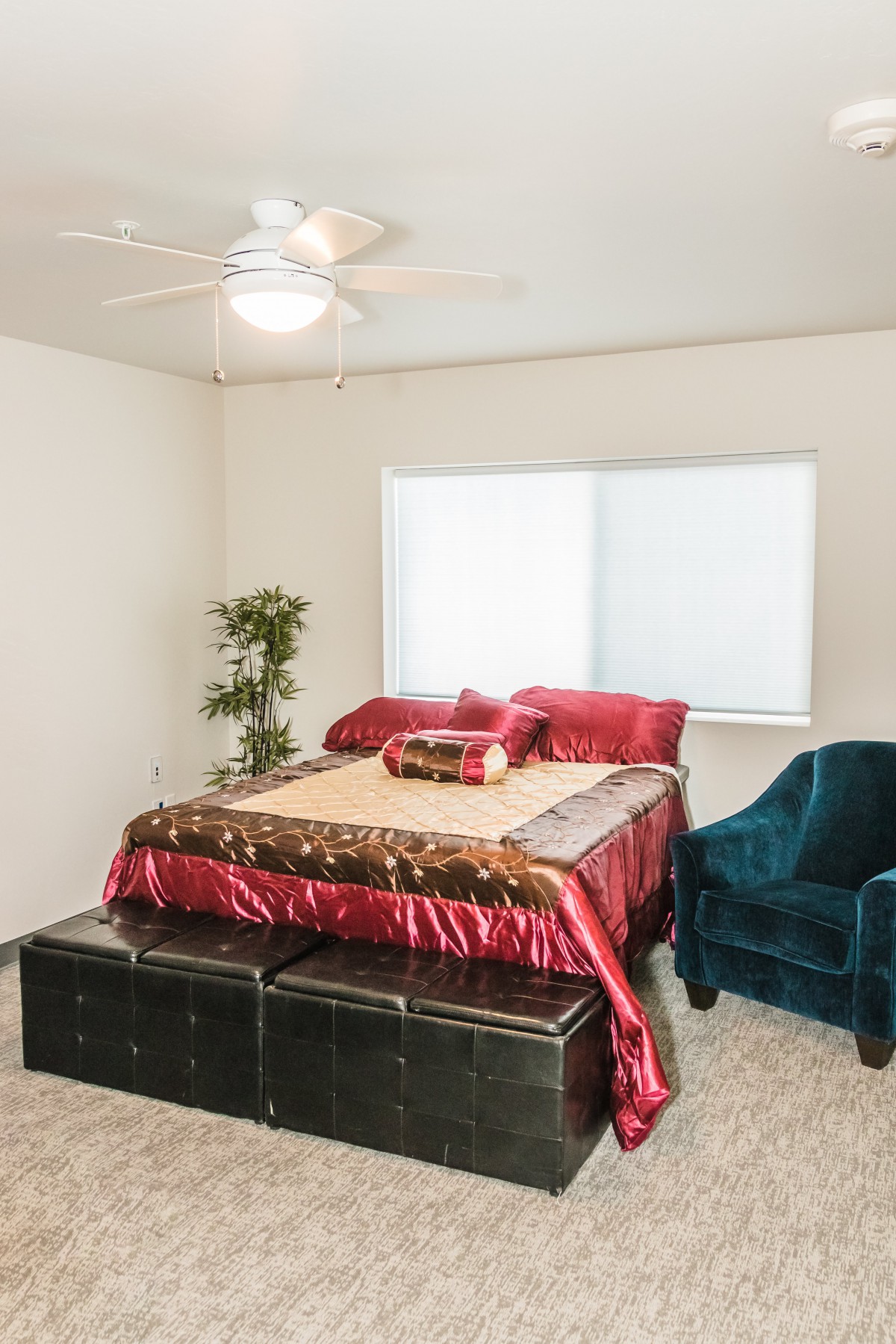 A cozy bedroom with red bedding, a teal armchair, and a potted plant near the window.