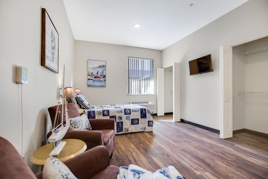 A modern hotel bedroom with a queen bed, armchairs, and a wall-mounted television.