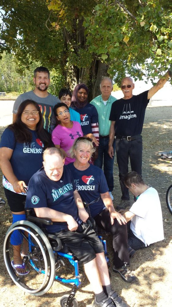 A diverse group of people gathered outdoors under a tree, with some wearing matching t-shirts and one person in a wheelchair.
