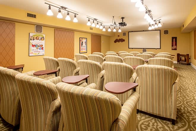 A cozy small theater room with multiple rows of plush armchairs facing a large projection screen, illuminated by overhead lights.