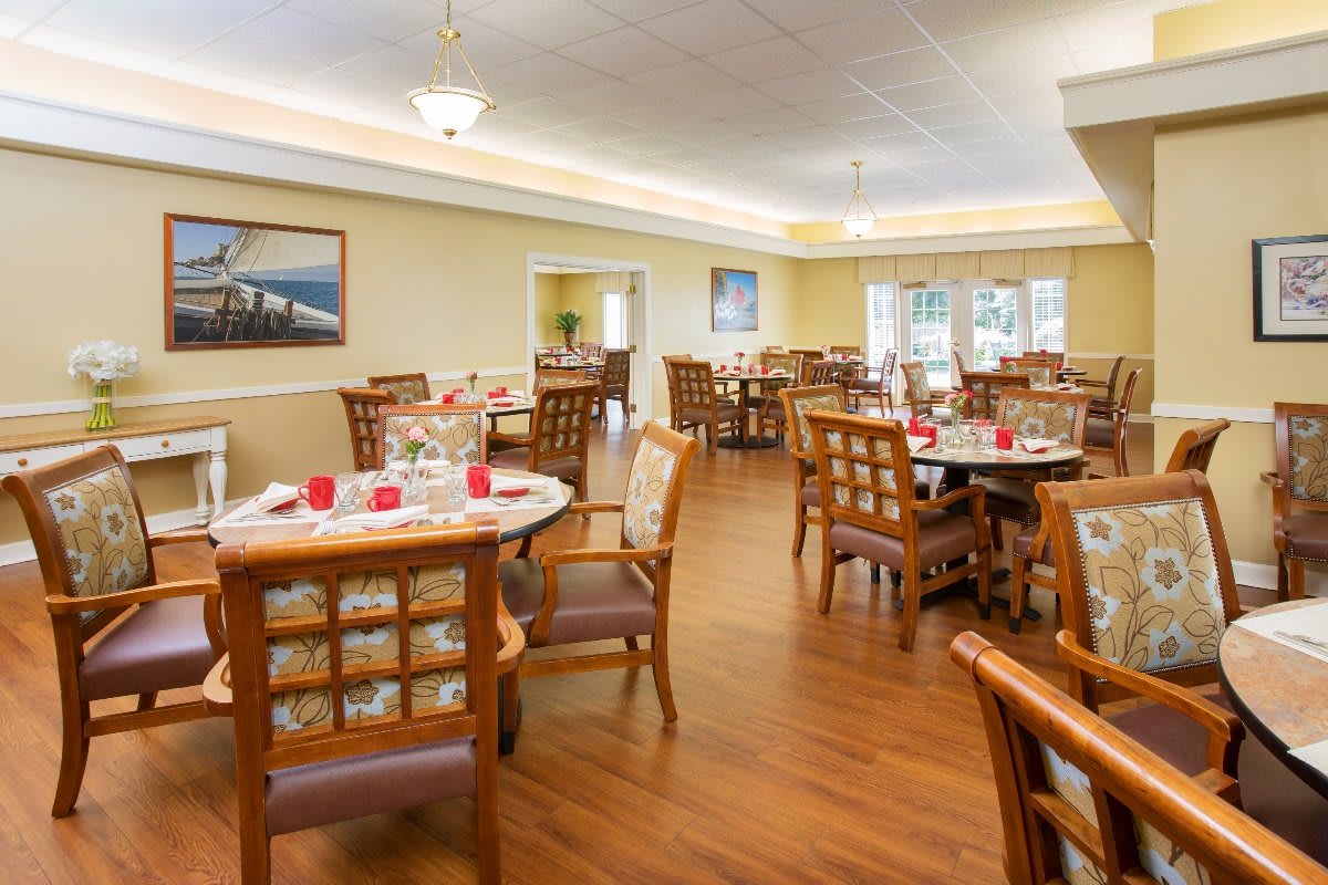A well-arranged dining room with neatly set tables and comfortable chairs, featuring warm lighting and wood flooring.