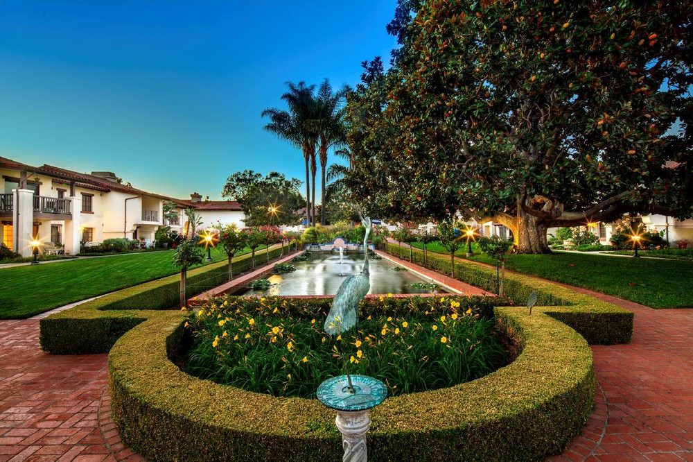 A beautifully landscaped garden with a central fountain surrounded by hedges and flowers, set against an evening sky.