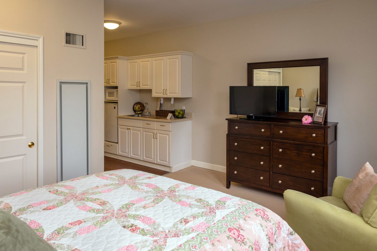 A cozy room with a quilted bedspread, dresser, and kitchenette.