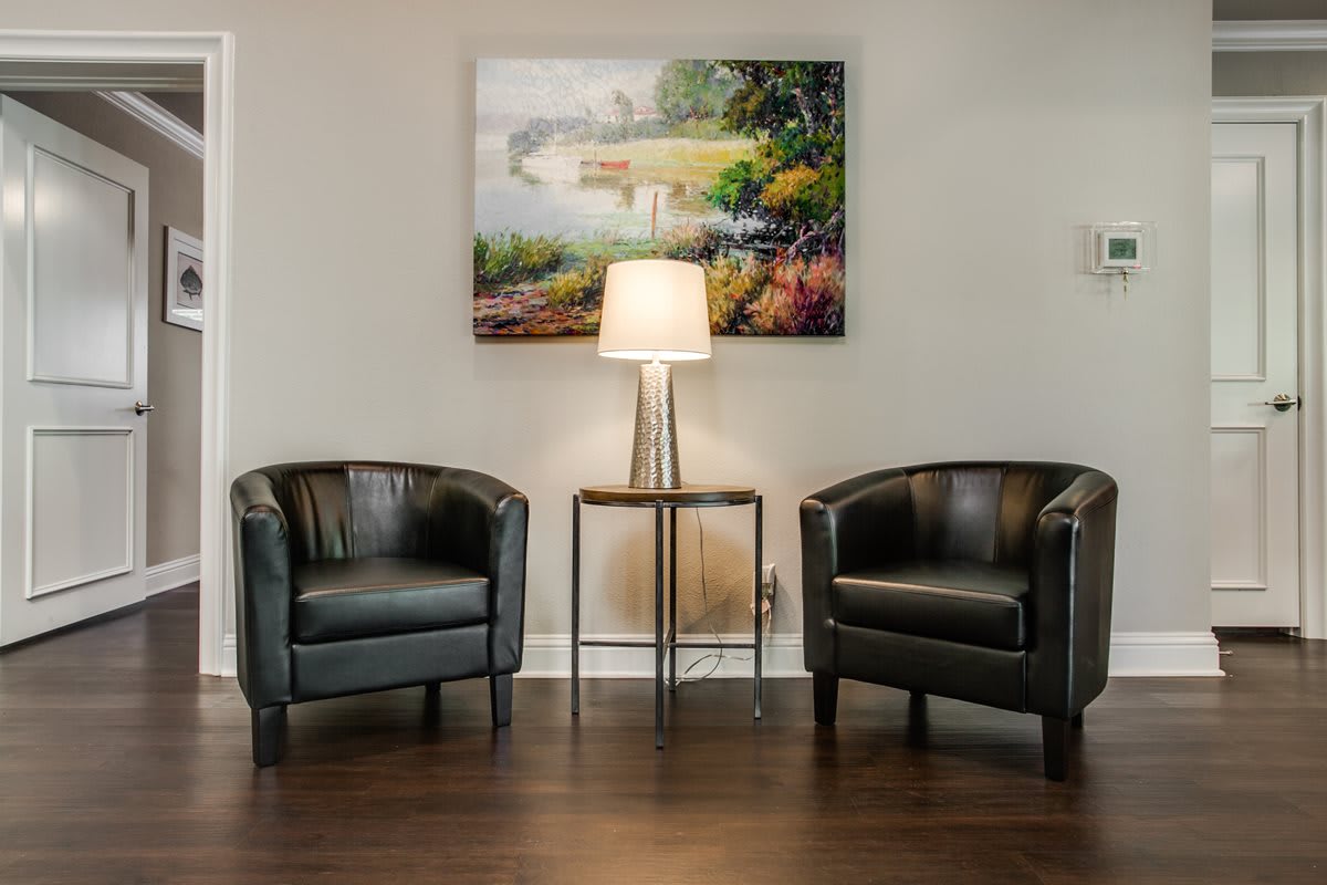 A cozy seating area with two black chairs, a lamp on a small table, and a landscape painting above.