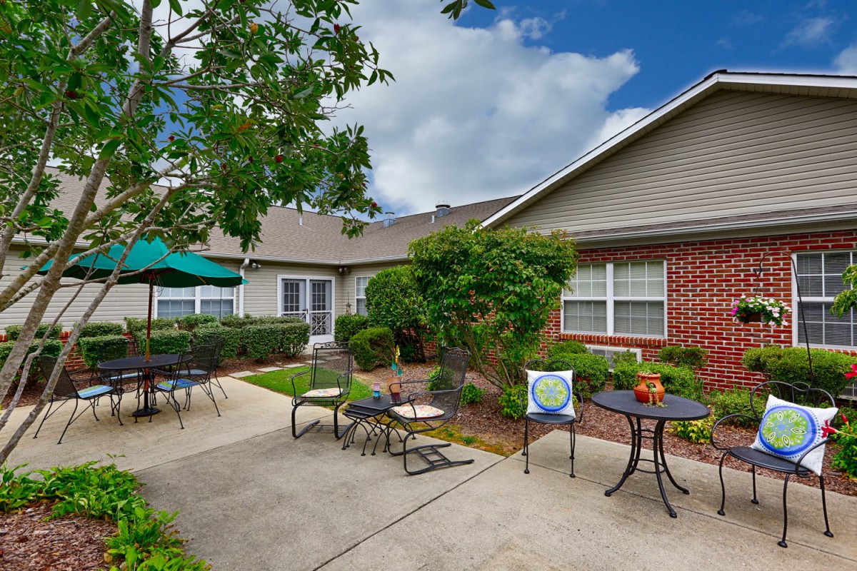 A cozy outdoor patio area with tables, chairs, and a sun umbrella, surrounded by greenery and adjacent to a building.