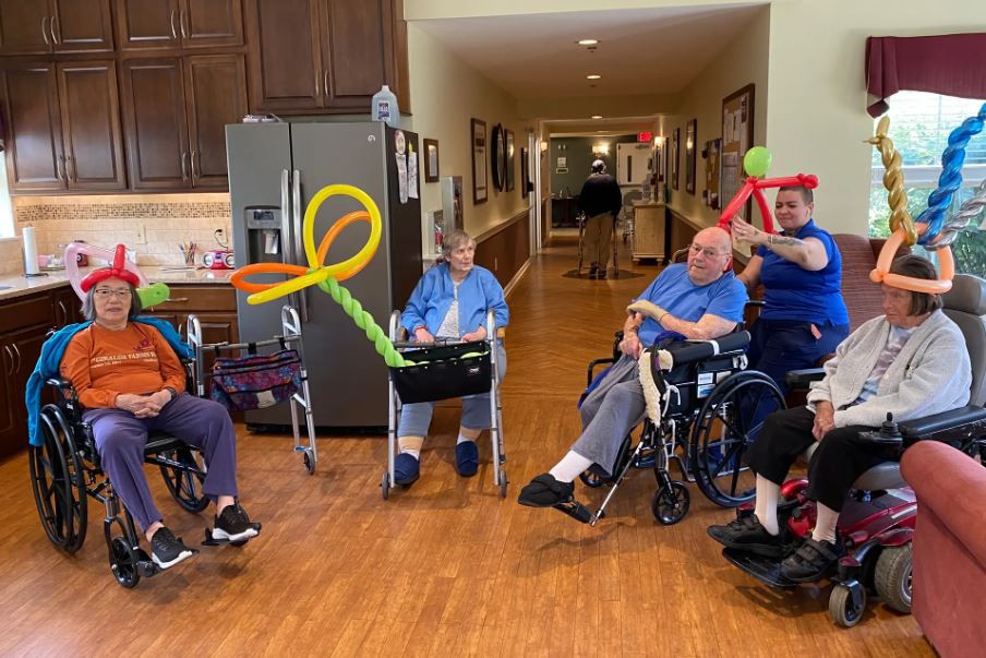 A group of elderly individuals in wheelchairs enjoying balloon creations in a care facility.