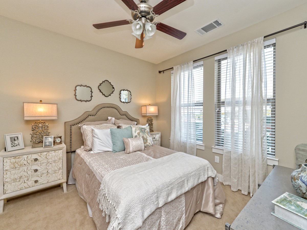 A cozy and elegantly decorated bedroom featuring a neatly made bed, two bedside lamps, and large windows with sheer curtains.