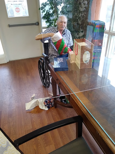 An elderly person in a wheelchair sits at a table surrounded by holiday gifts.