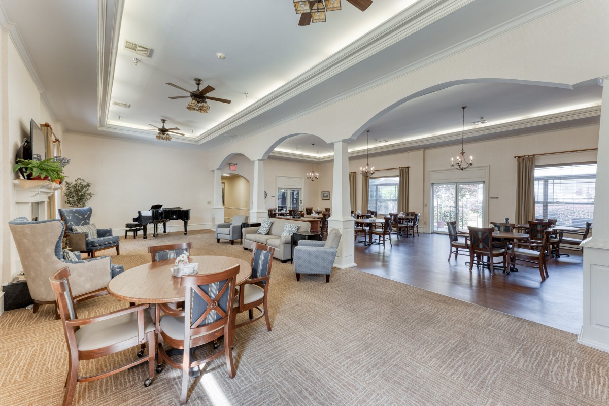 A spacious, elegantly designed dining area with round tables, chairs, a piano, and ceiling fans.