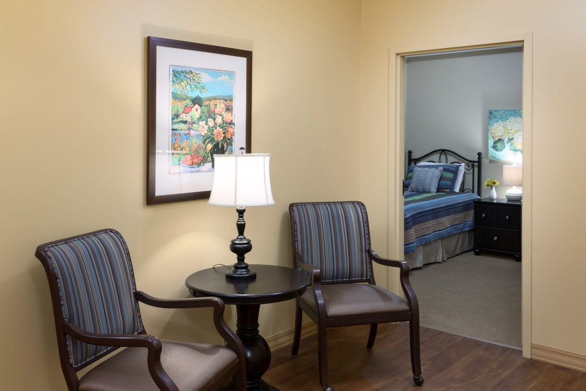 A cozy living area with two chairs, a round table, and a lamp, adjacent to a bedroom.
