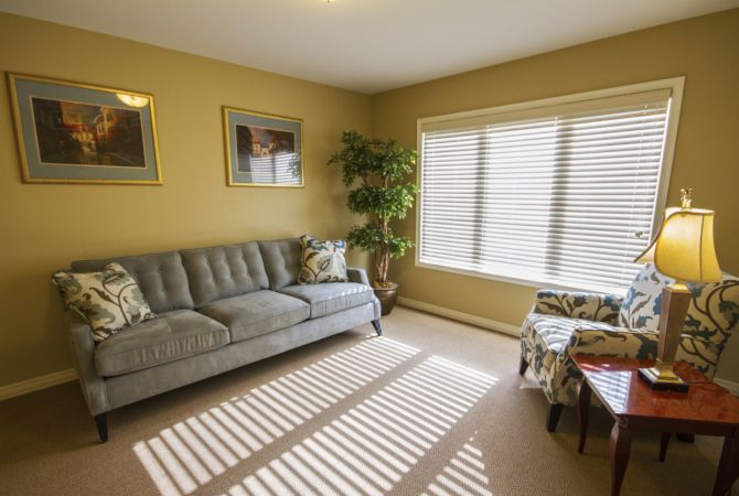 A cozy living room with a sofa, armchair, and large window with blinds filtering sunlight.
