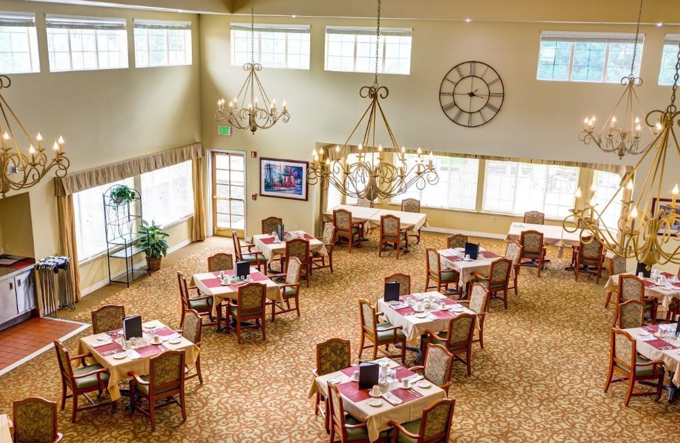 A spacious dining room with multiple tables, chairs, and chandeliers, featuring large windows that let in natural light.
