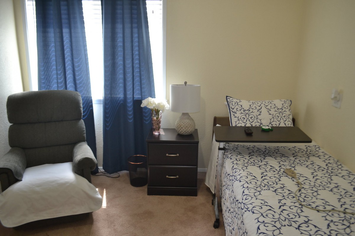 A cozy bedroom with a bed, nightstand, lamp, and chair by the window with blue curtains.
