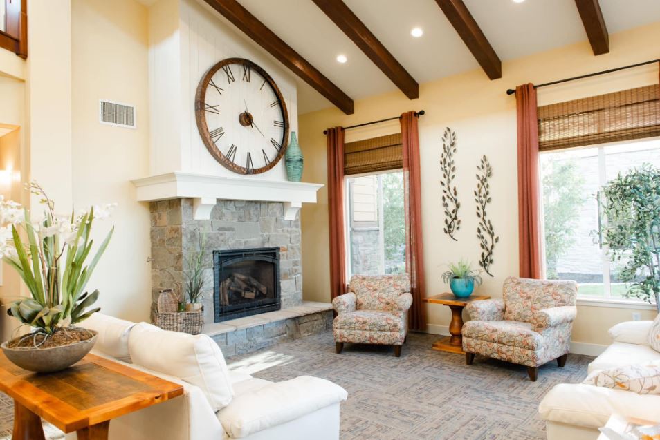 A cozy living room featuring a stone fireplace and a large wall clock, complemented by comfortable armchairs and wooden ceiling beams.