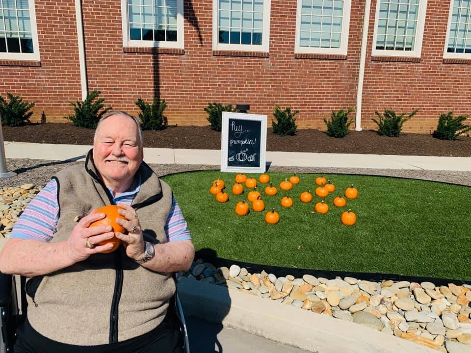A person smiling and holding a small pumpkin in front of a display of pumpkins on a grassy area outside a brick building.