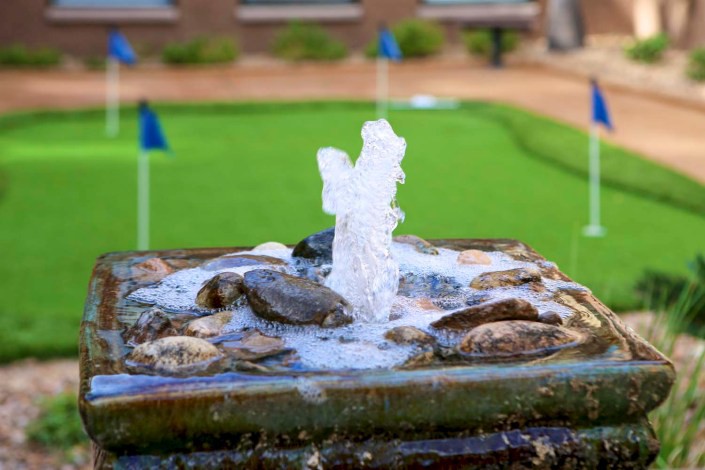 A small fountain with water flowing over rocks in front of a putting green with several flags.