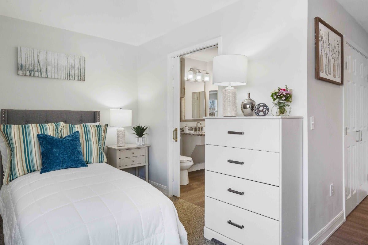 A cozy bedroom with a white dresser, bed, and nightstand, leading to an adjoining bathroom.