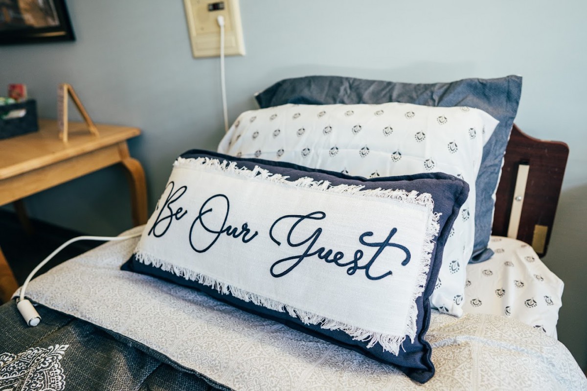 A cozy bed with a decorative pillow featuring the text 'Be Our Guest'.