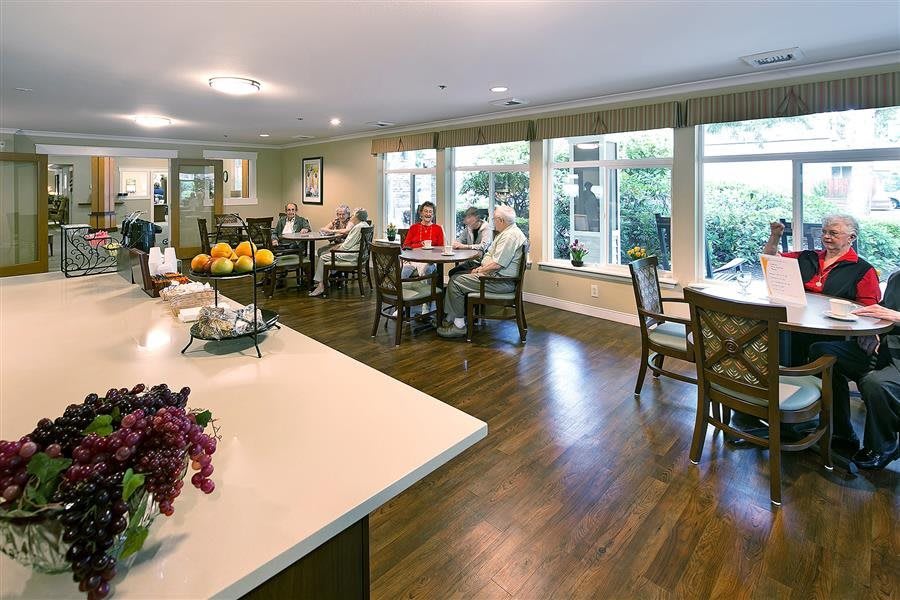 A communal dining space with tables and chairs, where several seniors are seated and enjoying a conversation.