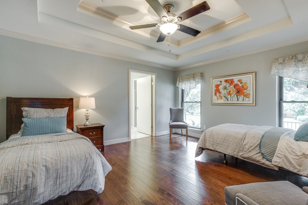 A spacious bedroom with two beds, wooden flooring, a ceiling fan, and a nightstand with a lamp beside the bed.
