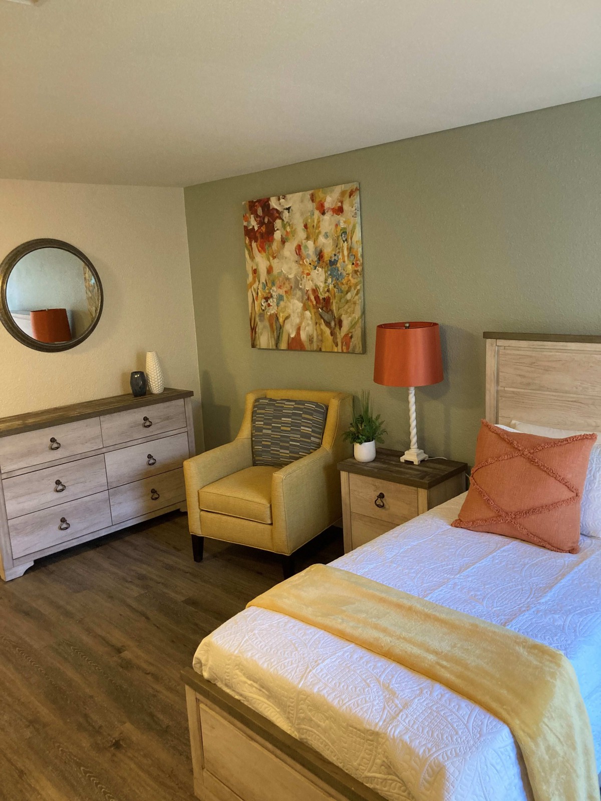 A cozy bedroom with modern decor, featuring a bed, an accent chair, a dresser, and colorful wall art.