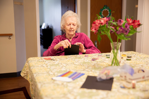 An elderly woman engaged in a crafting activity at a table adorned with flowers.