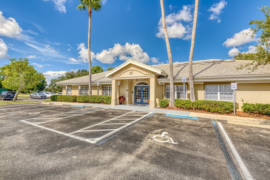 A single-story building with a parking lot and palm trees under a cloudy sky.