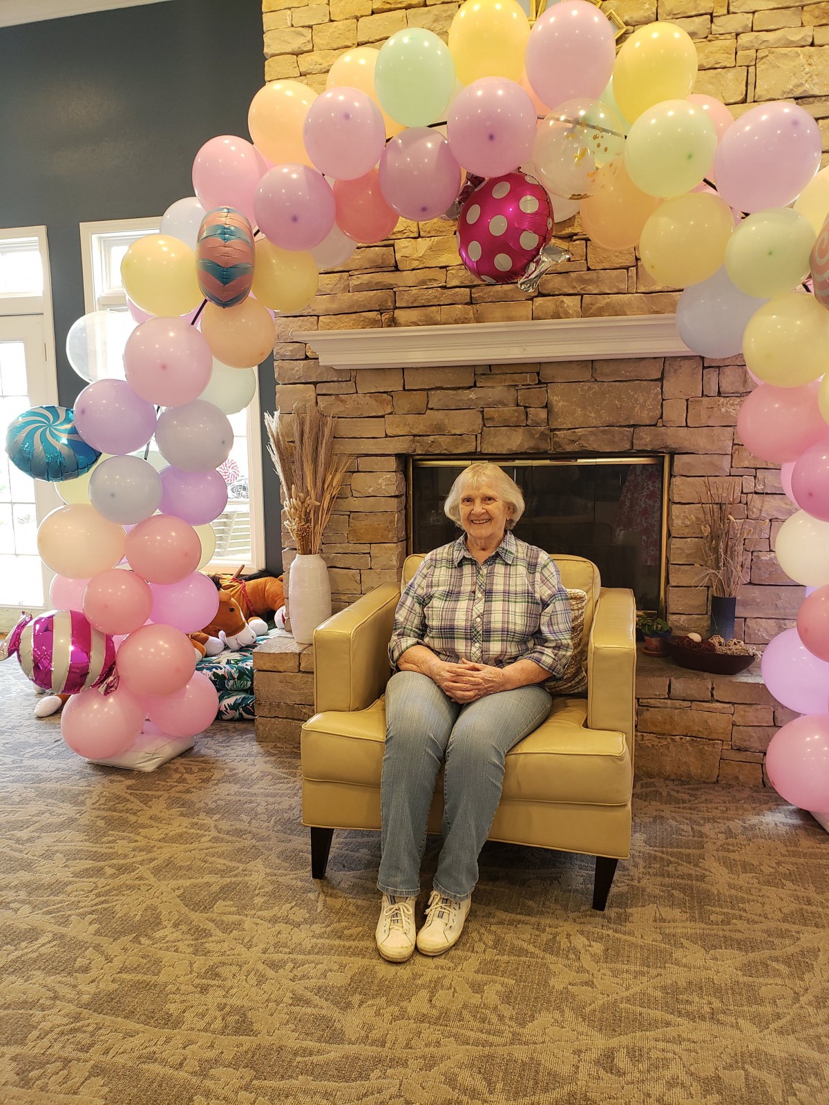 A person sitting on a chair surrounded by festive balloons in front of a fireplace.