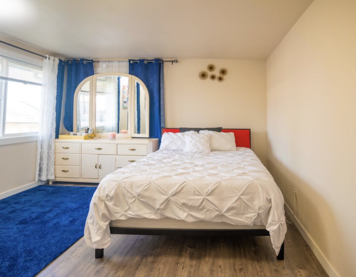 A cozy bedroom featuring a bed with white linens, blue curtains, and a dresser with a mirror.
