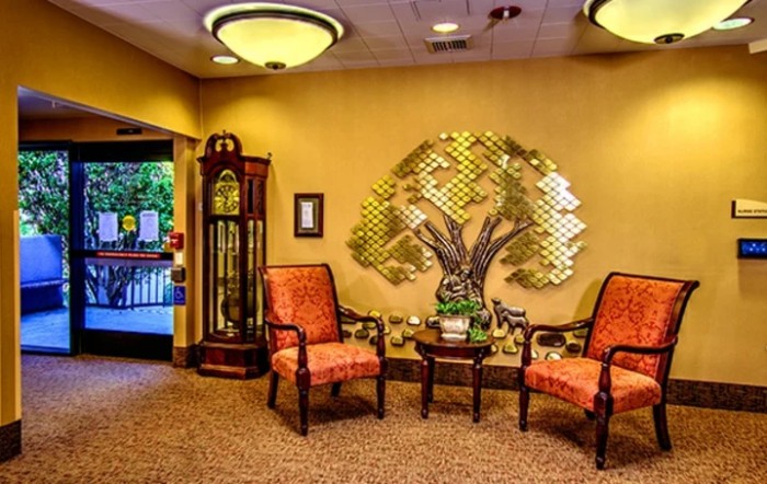 A warmly decorated lobby with two chairs, a table, a grandfather clock, and metal tree wall art.