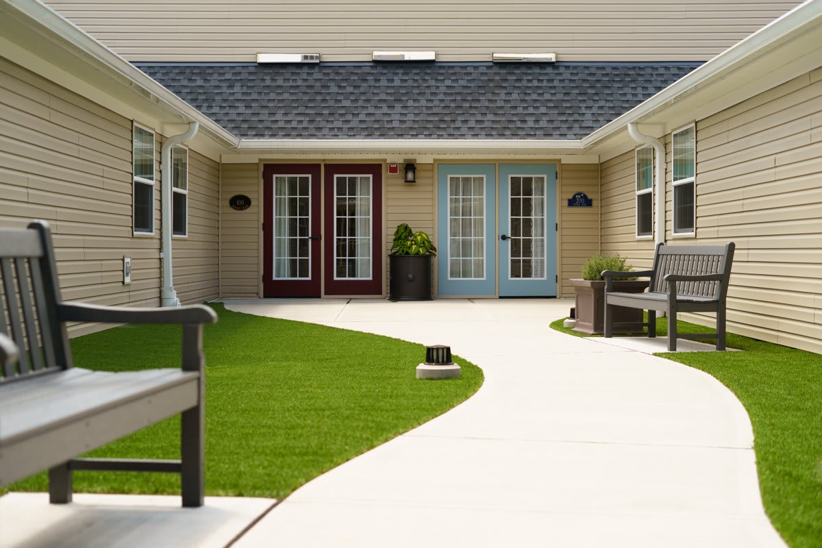 A neatly landscaped courtyard with benches, grass, and a smooth pathway leading to multiple doors of a building.
