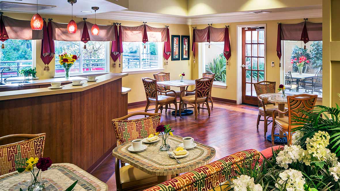 A cozy cafe interior with tables, chairs, and a serving counter, featuring large windows and warm decor.