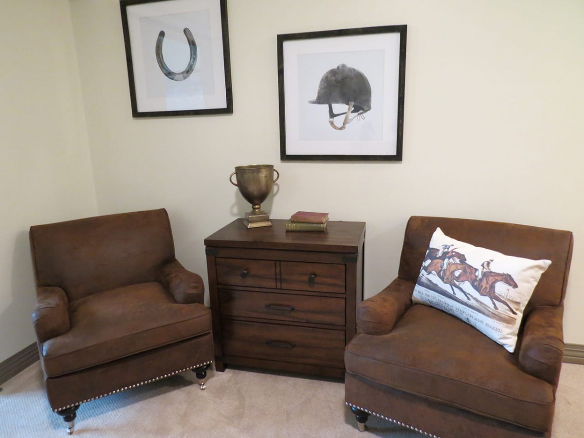 A cozy corner features two brown chairs, a wooden cabinet with a trophy on top, and equestrian-themed artwork on the walls.