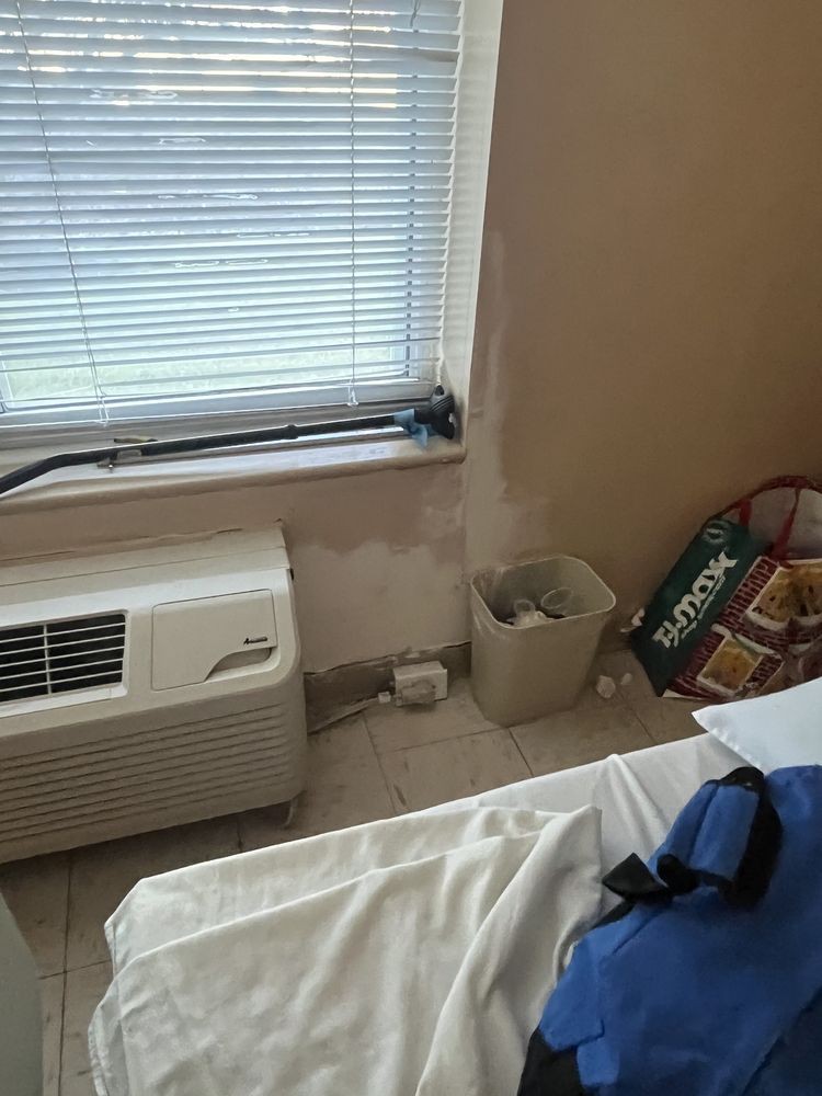 A hotel room corner with an air conditioner under a window, a trash can, snacks, and a partially visible bed.