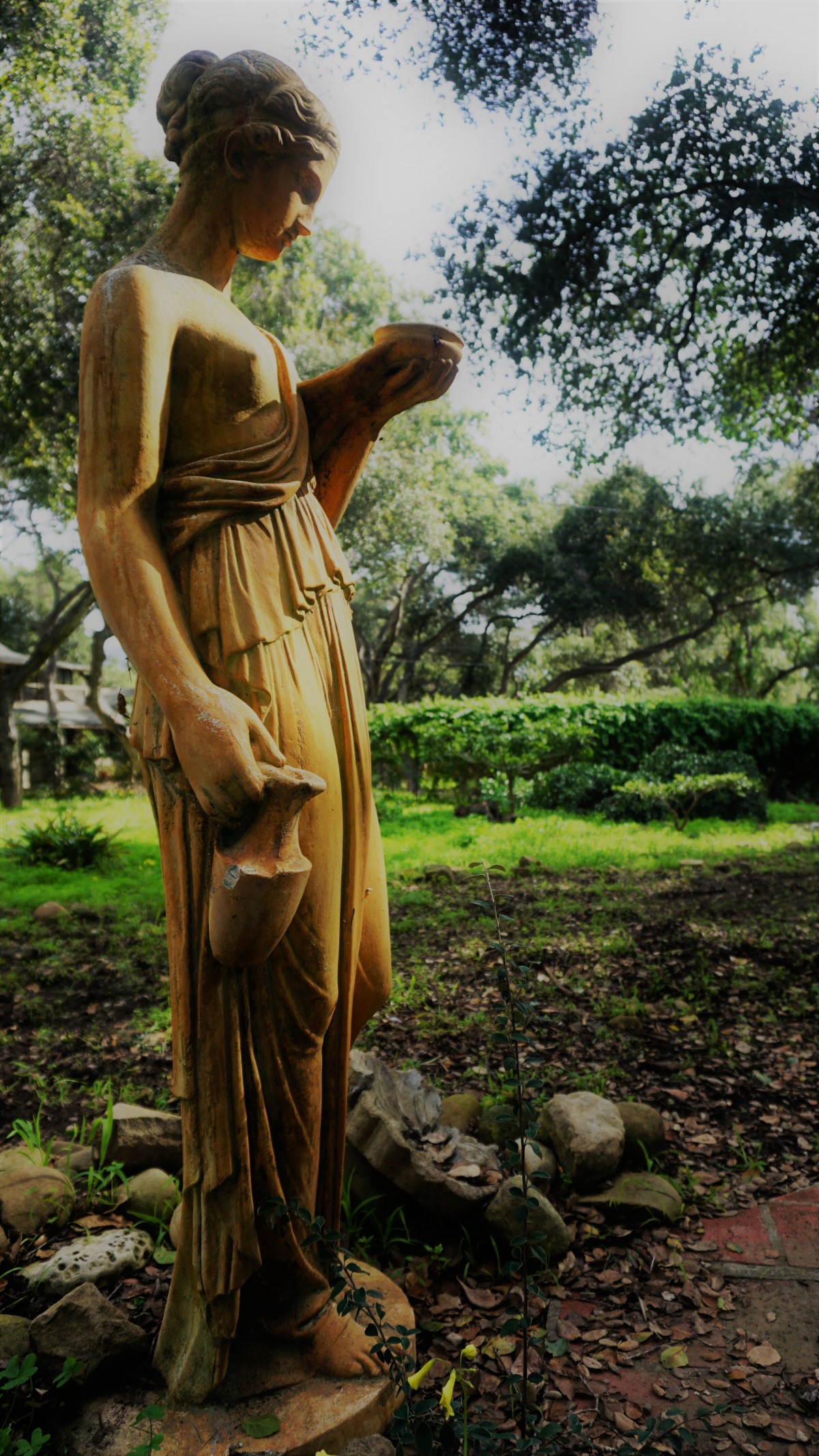 A classical statue in a serene garden setting surrounded by lush greenery and trees.