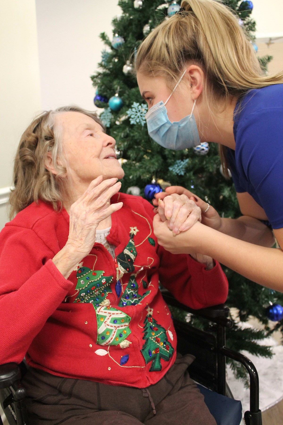 An elderly woman in a festive sweater shares a joyful moment with a masked healthcare worker beside a Christmas tree.