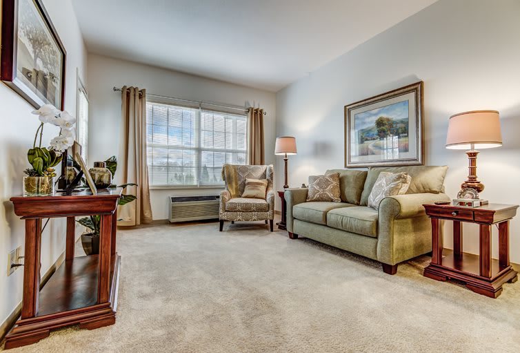 A cozy living room with a sofa, armchair, and soft carpet, complemented by a large window and decorative artwork.