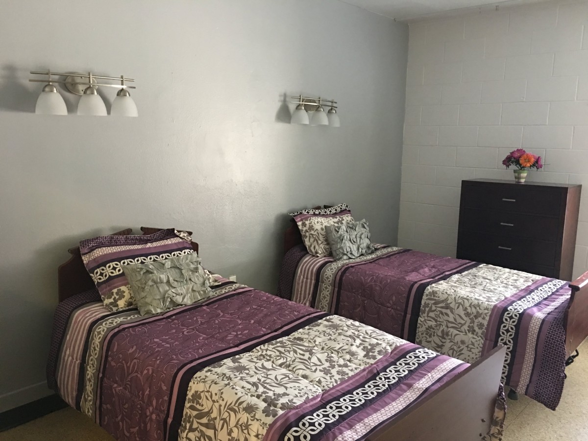 A bedroom featuring two twin beds with patterned bedding, wall-mounted lamps, and a dark dresser.