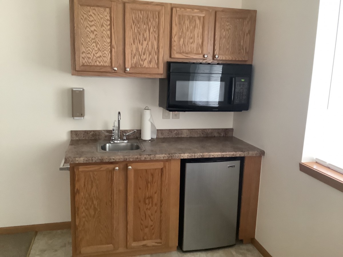 A compact kitchen setup with wooden cabinets, a microwave, mini fridge, and sink.