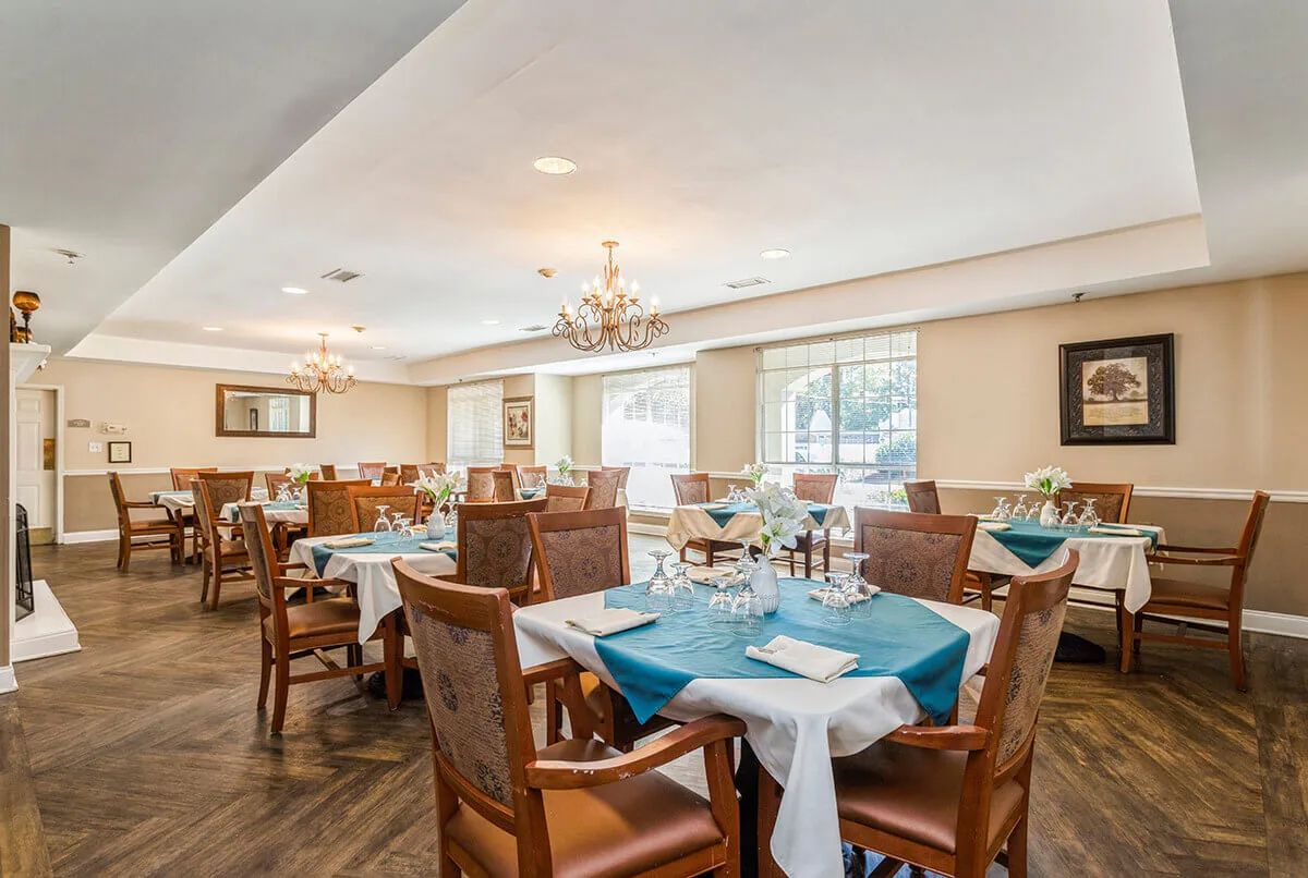 A well-decorated dining room with neatly arranged tables, each adorned with teal tablecloths and elegant place settings, under a large chandelier.