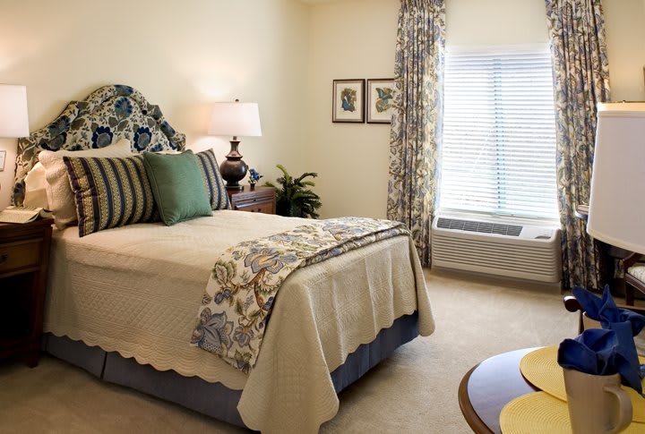 A cozy and well-decorated bedroom featuring a bed with patterned pillows, a lamp on the bedside table, and a window with curtains allowing natural light to brighten the space.