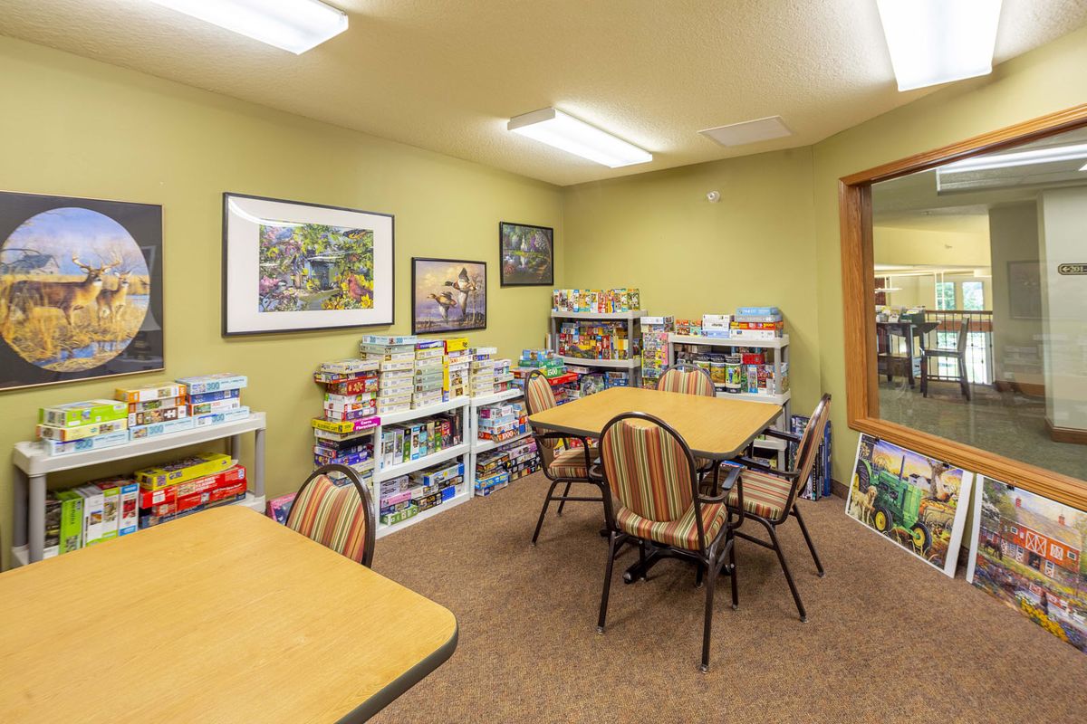 A cozy room with tables and chairs, surrounded by shelves filled with books and games, under bright lighting.