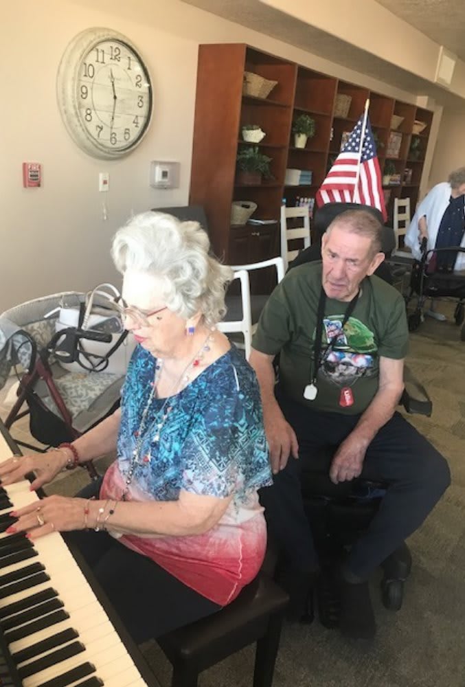 An elderly woman plays the piano while a man sits nearby, with a flag visible in the background.