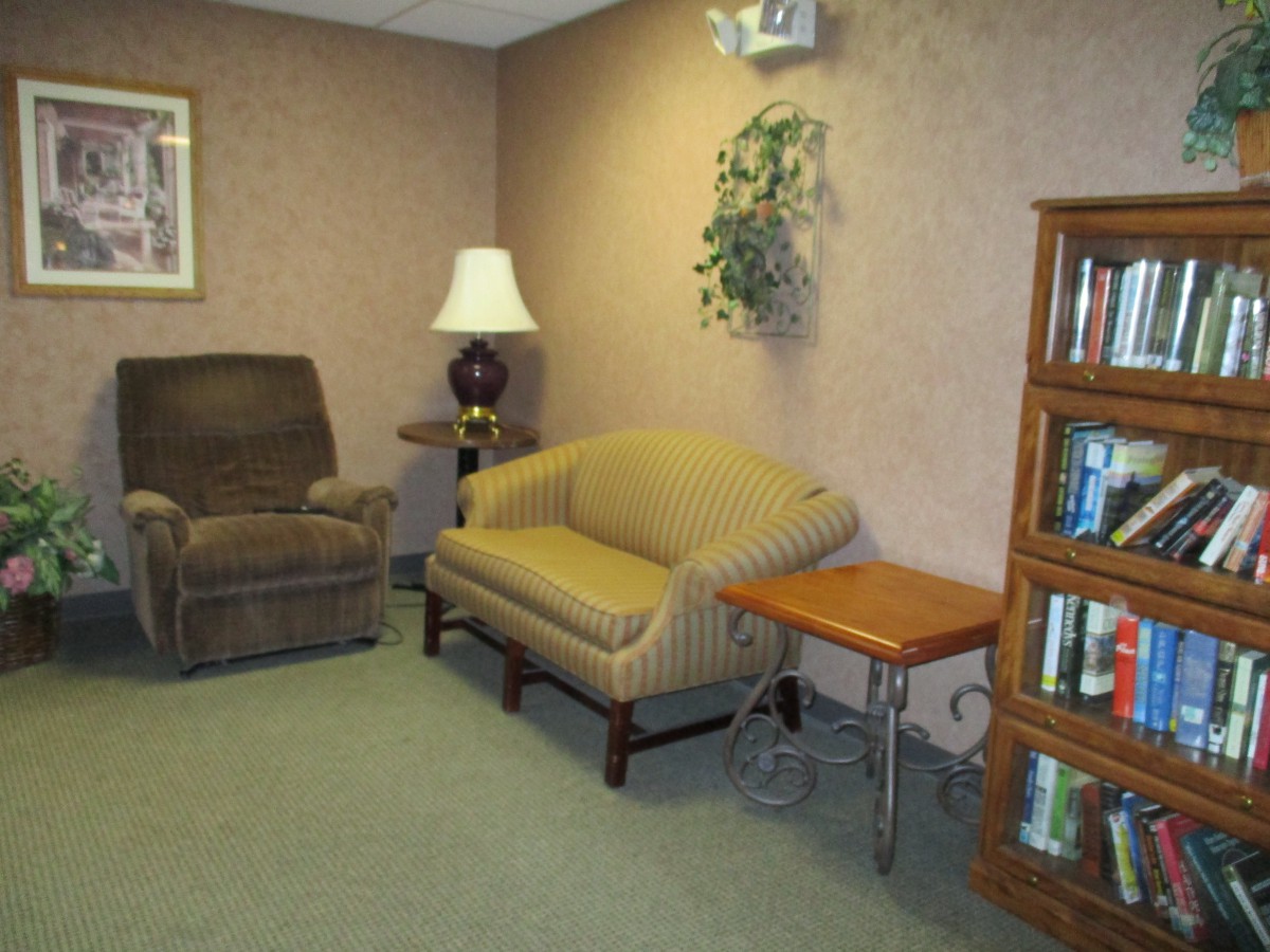 A cozy living room with an armchair, sofa, lamp, and a wooden bookshelf filled with books.