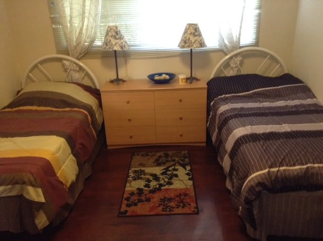 A cozy bedroom with twin beds, a central dresser topped with lamps, and a small rug on the floor.