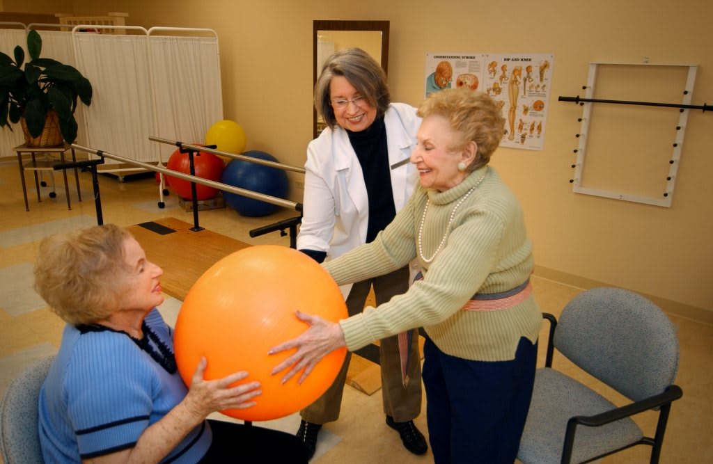 An elderly woman engages in a physical therapy session with healthcare professionals, using an exercise ball.