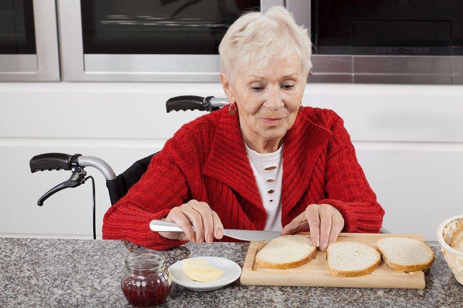An elderly woman in a red sweater is preparing a meal, spreading butter on slices of bread at a kitchen counter.