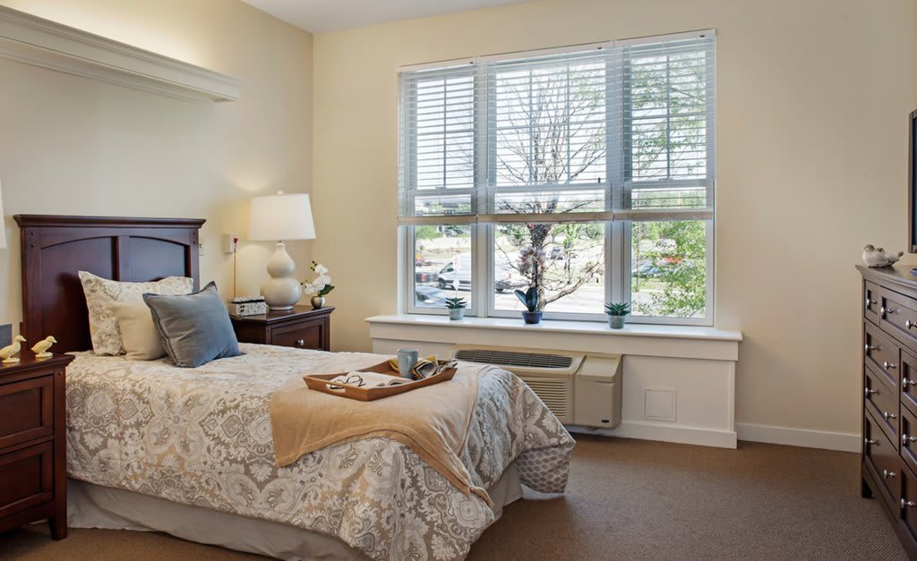 A cozy bedroom with a neatly made bed, illuminated by natural light from a large window.