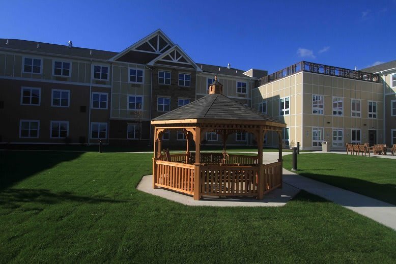 A wooden gazebo on a lush green lawn in front of a residential building under a clear blue sky.