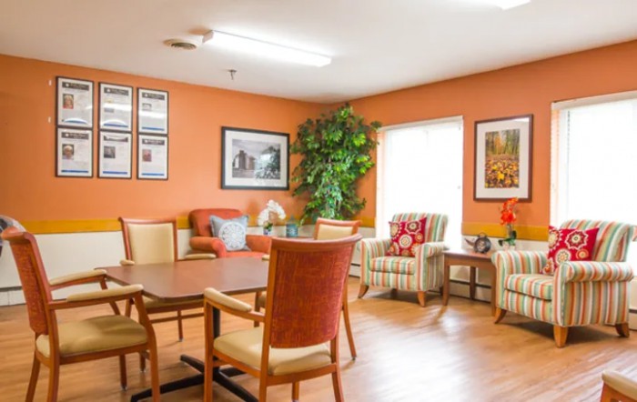 A cozy living room with orange walls, featuring a table surrounded by chairs, artwork on the walls, and a large plant by the window.